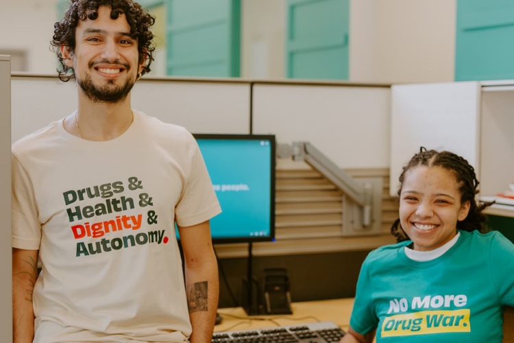 Young man wearing a shirt that reads "Drugs and Health and Dignity and Autonomy" and young Black woman wearing a shirt that reads "No More Drug War" sit in an office space smiling at the camera.