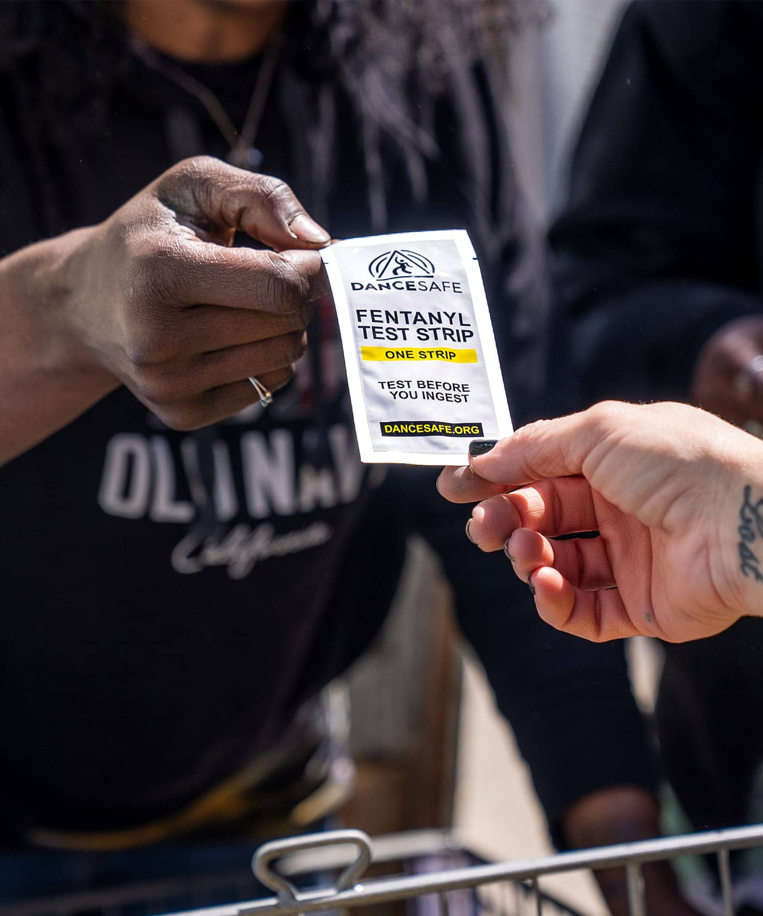 Photo someone being handed a small tear-open package that says "DanceSafe Fentanyl Test Strip. One Strip. Test before you inject. dancesafe.org"