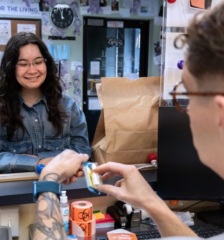 Photo of two people in a reception area. One person is behind the desk with their back to the camera holding naloxone. The other is looking through the reception window, smiling as if learning about naloxone.