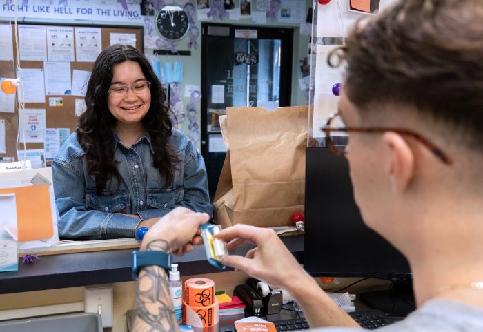 Photo of two people in a reception area. One person is behind the desk with their back to the camera holding naloxone. The other is looking through the reception window, smiling as if learning about naloxone.